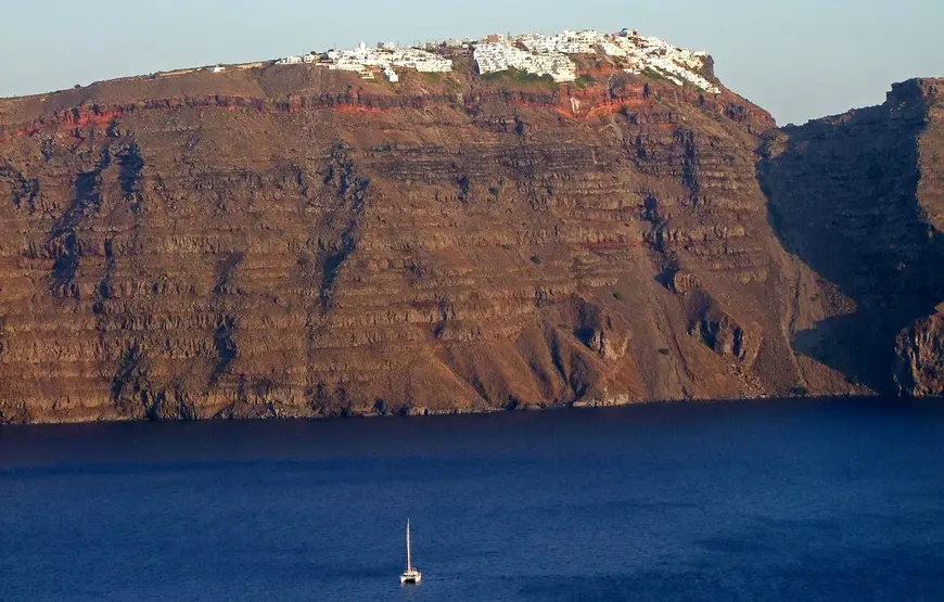 vista-caldera-di-santorini-roccia-rossa-mare-blu-sopra-i-villaggi-bianchi-e-sotto-una-barca-a-vela