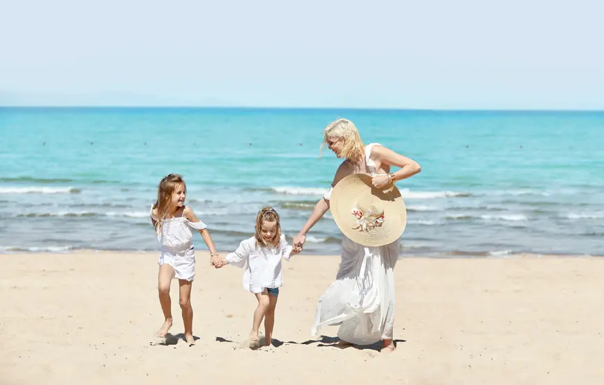mamma con due bambine. sulla-spiaggia-si-danno-la-mano-sono-vestite-di-bianco-alle-spalle-il-mare-azzurro